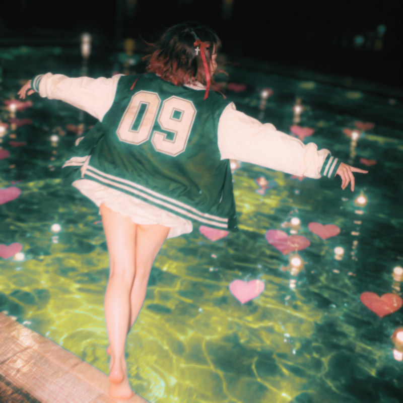 Judea — woman in a green varsity jacket numbered 09 walking the edge of a lit pool at night among floating pink hearts
